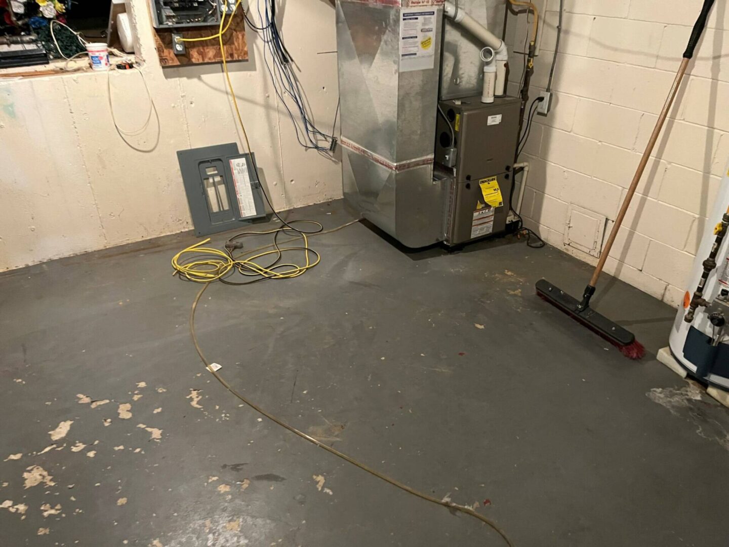 Basement with a furnace, electrical panel on the floor, broom, exposed pipes, and tangled cords. Walls are unpainted, and floor is bare concrete.