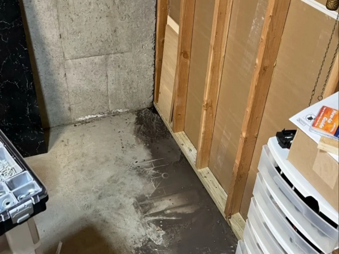 Unfinished basement corner under renovation by RAM Restoration, showing exposed studs, patched concrete floor, and nearby plastic drawers and toolbox.